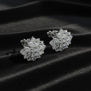 Könnte beinhalten: Ein Paar silberfarbene Ohrstecker. Jeder Ohrring hat eine Ansammlung kleiner, klarer Edelsteine in einem floralen Design. Die Ohrringe sind vor einem dunklen, strukturierten Hintergrund platziert, der ihr Funkeln hervorhebt.