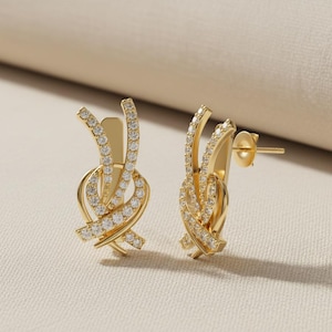 Könnte beinhalten: Ein Paar goldfarbene Ohrstecker. Jeder Ohrring hat ein Knotendesign mit gewölbter Oberseite und ist mit kleinen, funkelnden, klaren Steinen besetzt. Die Ohrringe haben einen Stecker und einen Butterfly-Verschluss.