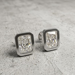 May include: A pair of square-cut diamond stud earrings. Each earring has a rectangular, clear gemstone set in a silver bezel. The earrings are shown on a textured, grey surface.