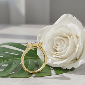 May include: A gold engagement ring with a large, clear gemstone is displayed on a green leaf next to a white rose. The ring has a detailed band with smaller stones. The setting is a classic design.