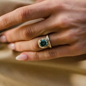 Puede incluir: Un anillo de oro con una piedra preciosa cuadrada de color verde azulado. El anillo está engastado en un bisel de oro y tiene una banda sencilla y elegante. El anillo se muestra en una mano con un fondo neutro. Es una joya.
