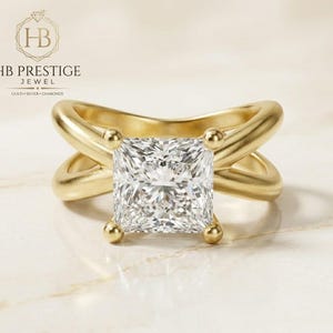Pode incluir: Um anel de ouro com um grande diamante central de corte quadrado. A banda apresenta um design cruzado único. O anel está sobre um fundo neutro, com o texto "HB PRESTIGE JEWEL" visível no canto superior esquerdo.