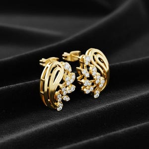 May include: A pair of gold stud earrings with a floral design, set with small, clear gemstones. The earrings are displayed on a black, textured surface, highlighting their intricate details and sparkling accents.