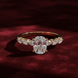 May include: A gold ring with a large oval-cut diamond center stone, flanked by smaller pear-shaped diamonds. The ring is set against a deep red background, highlighting the sparkle of the gemstones. The band is also adorned with small diamonds.