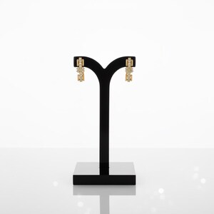 May include: A pair of gold-colored earrings with a rectangular design, adorned with small, sparkling stones. The earrings are displayed on a black jewelry stand against a white background. The earrings have a geometric shape.