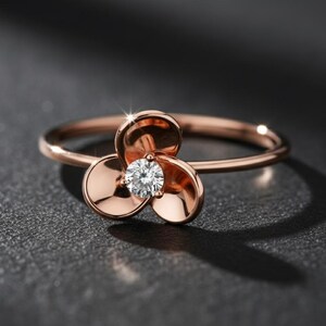 Puede incluir: Anillo de oro rosa con un diseño de flor de tres pétalos. En el centro, un diamante redondo y brillante. El anillo se muestra sobre un fondo oscuro y texturizado, resaltando su elegancia.