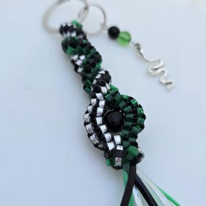 May include: A handmade keychain featuring a woven design in black, green, and silver beads. The keychain includes a silver key ring, a dangling charm with green and black beads, and a silver spiral charm. Black, silver, and green ribbons are attached.