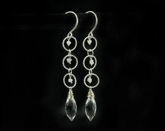 May include: Silver dangle earrings with clear teardrop-shaped crystals and small white beads hanging from three silver circles on each earring.