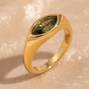 Może przedstawiać: Złoty pierścionek z zielonym kamieniem szlachetnym o szlifie markiza. Pierścionek ma gładką, wypolerowaną obrączkę i oprawę w kształcie ramki, która bezpiecznie trzyma kamień. Kamień ma jasnozielony kolor.