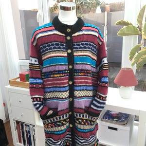 Könnte beinhalten: Ein langer, mehrfarbiger Strickcardigan mit schwarzem Kragen und Besatz. Der Pullover hat horizontale Streifen in Rot-, Blau-, Lila- und Cremetönen mit dekorativen Mustern und Taschen. Er ist zugeknöpft und auf einer Schaufensterpuppe ausgestellt.