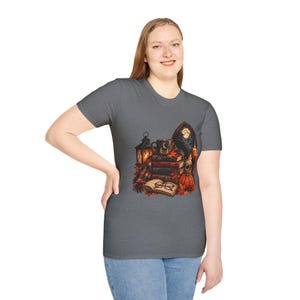 May include: A dark gray short-sleeve t-shirt featuring a graphic print of a stack of books, a lantern, a mug, a raven, a skull, a pumpkin, and an open book with glasses. The design includes autumn leaves and a moonlit window background.