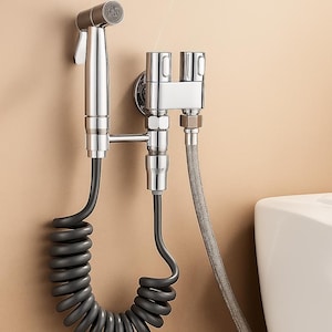 Peut inclure: Un pulvérisateur de bidet chromé avec un tuyau noir enroulé et une vanne de commande argentée. Le pulvérisateur est monté sur un mur beige près d'une toilette blanche. Le bidet est équipé d'une buse et d'un mécanisme de contrôle de l'eau.