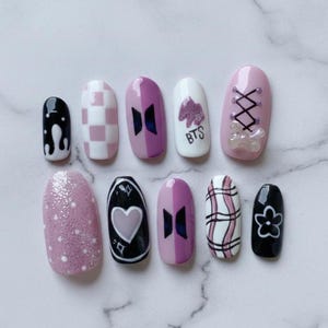 May include: A set of ten artificial nails with various designs. The nails feature a mix of colors including black, white, and shades of pink and purple. Some nails have checkered patterns, heart shapes, and the text "BTS".