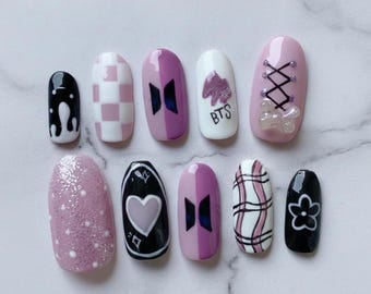 May include: A collection of ten artificial nails in various designs. The nails are painted in black, white, and pink and purple hues. Some nails feature checkered patterns, heart shapes, and the text "BTS".