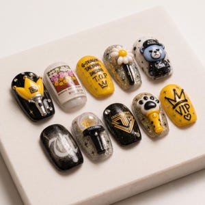 May include: A set of ten press-on nails featuring Big Bang themed designs. The collection includes black, yellow, and glitter finishes with 3D elements like a crown, a daisy, a bear, a paw print, and miniature light sticks with text.