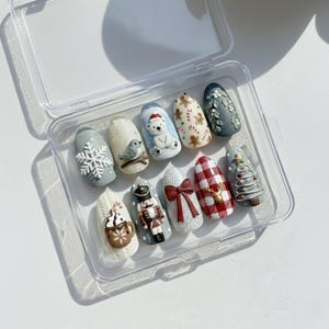 May include: A set of ten artificial nails with festive Christmas designs. Each nail features unique 3D art, including a snowflake, bird, polar bear, gingerbread men, a bow, a reindeer, and a Christmas tree. The nails are displayed in a clear plastic case.