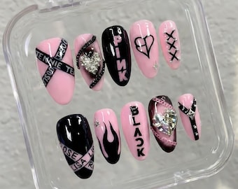 May include: A set of ten press-on nails in a clear plastic case, featuring a black and light pink color scheme. Designs include the text PINK and BLACK, heart shapes, flame patterns, cross-stitch details, and silver rhinestones on almond-shaped tips.