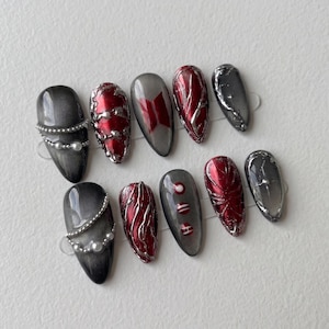 May include: A set of ten artificial nails in various designs. The nails feature a gradient gray base with silver and red accents. Some nails have pearl embellishments or a logo. The nail shapes are oval.