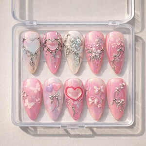 May include: A set of ten artificial nails in a clear plastic case. Each nail is a different design, featuring pink and white bases with silver embellishments of hearts, flowers, and butterflies. Some nails have glitter accents.
