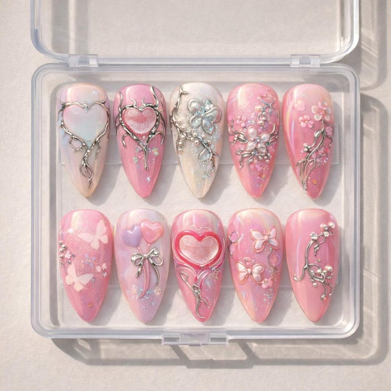 May include: A set of ten artificial nails in a clear plastic case. Each nail is a different design, featuring pink and white bases with silver embellishments of hearts, flowers, and butterflies. Some nails have glitter accents.