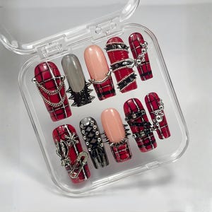 May include: A set of artificial nails in a clear plastic case. The nails feature a variety of designs, including red plaid, solid gray, and nude pink. Some nails are adorned with silver chains, spikes, and other metal embellishments.