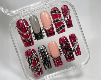 May include: A set of artificial nails in a clear plastic case. The nails feature a variety of designs, including red plaid, solid gray, and nude pink. Some nails are adorned with silver chains, spikes, and other metal embellishments.