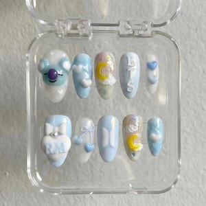 Puede incluir: Un juego de diez uñas postizas en una caja de plástico transparente. Las uñas están pintadas en azul pastel, blanco y beige con varios diseños, incluyendo un koala de dibujos animados, nubes, lunas, corazones y las letras "BTS" y "RM".