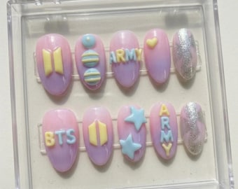 Könnte beinhalten: Ein Set aus zehn oval geformten Press-on-Nägeln in einer transparenten Kunststoffbox. Die Nägel weisen einen Farbverlauf aus Rosa und Lila mit verschiedenen Designs auf, darunter die Buchstaben "BTS" und "ARMY", Sterne und andere dekorative Elemente.