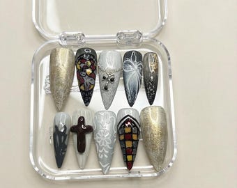 May include: A set of ten stiletto-shaped press-on nails in a clear plastic case. The designs include gold foil textures, stained glass patterns, white lace-like filigree, a silver cross charm, a dark brown 3D cross, and white swirling line art.