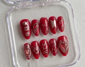 May include: A set of red press-on nails with silver accents. Designs include geometric shapes, swirls, a heart, and the letters "BTS". The nails are oval-shaped and displayed in a clear plastic case.