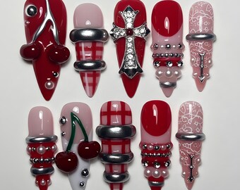 May include: A set of ten artificial nails in various designs. The nails feature a mix of red, pink, and white colors, with embellishments like cherries, silver accents, and pearl-like beads. Some nails have a cross design or a lattice pattern.