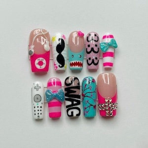 Könnte beinhalten: Ein Set aus zehn künstlichen Fingernägeln mit verschiedenen Designs. Die Designs umfassen einen rosa-weiß gestreiften Nagel mit einer Schleife, einen Nagel mit dem Wort "SWAG" und einen Nagel mit einem Fernbedienungsdesign. Weitere Nägel zeigen einen Schnurrbart, einen Hai und ein Blumenmuster.
