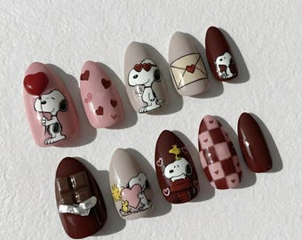 May include: A set of ten almond-shaped press-on nails with various designs. The nails feature Snoopy characters, hearts, and chocolate bar accents in shades of pink, brown, and beige. Some nails have 3D elements, like a heart balloon.