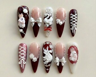 May include: A set of ten long, almond-shaped artificial nails. Designs include white bows, roses, swans, and a rabbit. Colors include burgundy, pink, and white, with pearl and rhinestone accents. The nails are arranged on a light-colored surface.