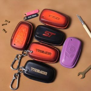 Puede incluir: Una colección de fundas de cuero para llaveros en varios colores, incluyendo rojo, negro y morado. Algunas fundas presentan texto en relieve como "F-150", "ST" y "TREMOR". Una funda morada tiene un diseño de girasol. También hay llaveros y herramientas.