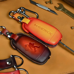 May include: Two leather key fob cases, one orange with "MAVERICK" embossed, the other brown with a Bigfoot design and American flag. Both have key rings and are on a brown leather surface.