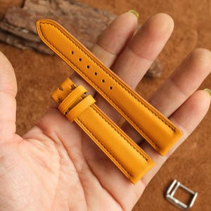 May include: A pair of bright yellow leather watch straps with matching stitching and a buckle. The straps are held in a hand against a brown background. The straps are new and unused.