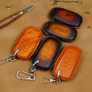 May include: A collection of handcrafted leather key fob covers in various shades of brown and orange. Some covers feature embossed designs, including the text "F150" and an American flag. Each cover has a metal keyring.