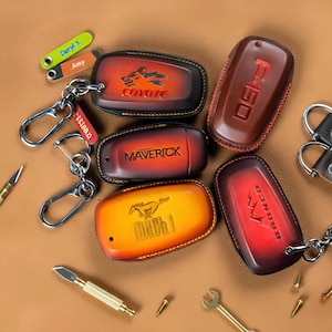 May include: A collection of leather key fob covers in various colors, including brown, orange, and red. Each cover features a different vehicle name or logo, such as "Coyote," "Maverick," and "Bronco." Keychains and tools are also visible.