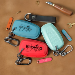 Puede incluir: Una colección de llaveros y fundas para llaves en varios colores, incluyendo azul, rojo y verde menta. Las fundas para llaves tienen el texto "Bronco Babe" impreso. También se incluyen pequeñas etiquetas con nombres o iniciales.