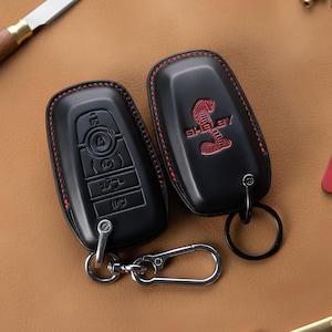 May include: Two black leather key fob covers with red stitching. One has a key fob with buttons, the other has a red Shelby logo. Both have key rings and a silver carabiner clip.