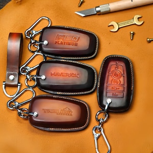 May include: A collection of brown leather key fobs with silver key rings. The fobs are embossed with the names "F150 PLATINUM", "MAVERICK", and "TREMOR OFFROAD". A matching leather strap is also included. The background is a tan leather surface.