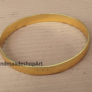 May include: A gold-colored bangle bracelet with a hammered texture. The bracelet is circular and appears to be made of metal. The text "HandmaadeshopArt" is printed on the bracelet. The bangle is displayed on a light brown surface.