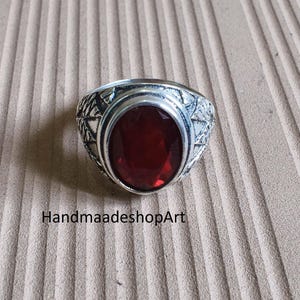 Puede incluir: Un anillo de plata con una piedra preciosa ovalada de color rojo intenso. El anillo tiene un diseño detallado y estampado alrededor de la piedra. El texto "HandmaadeshopArt" es visible.