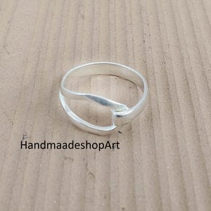May include: A silver ring with a unique design. The ring features a split band that merges into a single, slightly raised element. The text "HandmaadeshopArt" is visible on the surface.