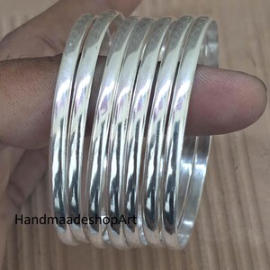Op de afbeelding: Een set van zes zilverkleurige armbanden met een glad, afgerond oppervlak. De armbanden zijn gestapeld gerangschikt en reflecteren het licht op hun metalen afwerking. De tekst HandmaadeshopArt is zichtbaar in de linkerbenedenhoek.