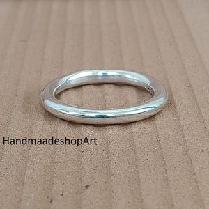 Pode incluir: Um anel de prata com um design liso e arredondado. O anel é feito de um metal brilhante e reflexivo e está sobre um fundo castanho. O texto "HandmaadeshopArt" é visível.