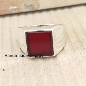 Può includere: Un anello d'argento con una gemma quadrata rosso scuro. L'anello ha una fascia larga e un design geometrico moderno. Il testo "HandmaadeshopArt" è visibile. Adatto per l'uso quotidiano o occasioni speciali.