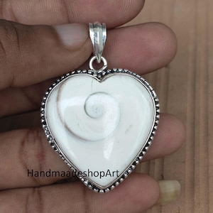 May include: A heart-shaped pendant featuring a smooth, off-white Shiva eye shell with a natural spiral pattern, encased in a polished silver-toned metal setting with a beaded border and a silver bail. The text HandmaadeshopArt is visible.