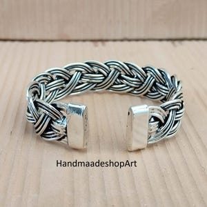 May include: A silver bracelet with a woven design. The bracelet features a braided pattern and rectangular clasps. The text "HandmaadeshopArt" is visible on the surface.